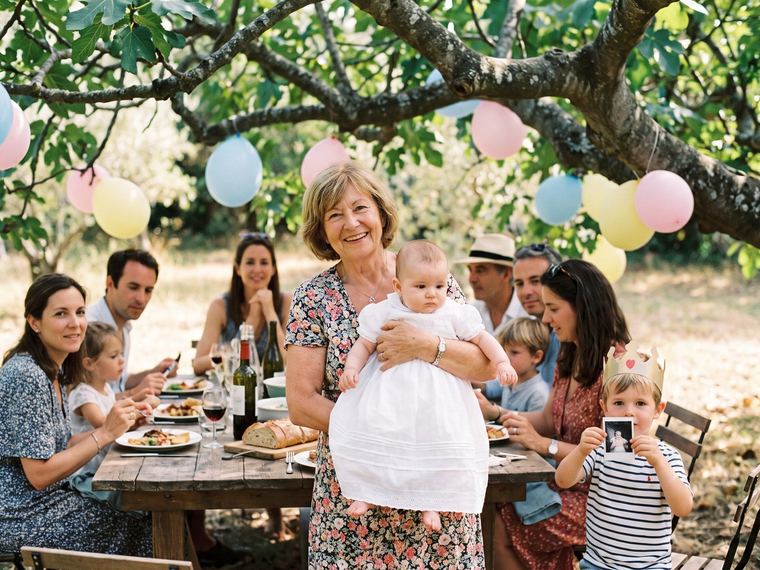 Famille réunie autour d’une table décorée de blanc dans un jardin provençal, un bébé dans les bras de sa grand-mère, pour un baptême au printemps