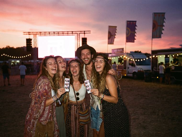 Cinq amis qui rient en brandissant leurs bandes de photos sur un site de festival au coucher du soleil, avec la scène, des oriflammes, des guirlandes lumineuses et des food trucks en arrière-plan