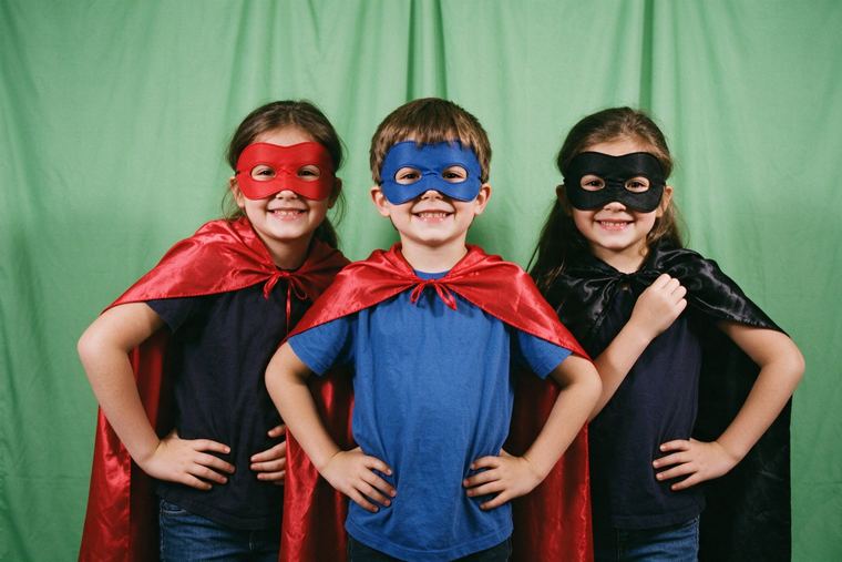 Trois enfants en capes de super-héros qui prennent la pose devant un fond vert, prêts pour l’incrustation