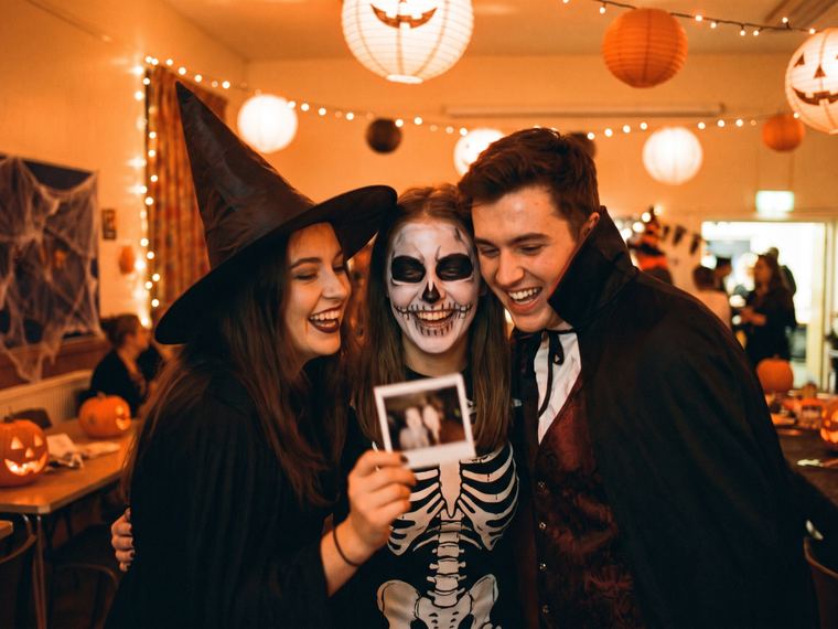 Amis déguisés en sorcière, squelette et vampire dans une salle décorée pour Halloween, l’une d’eux montre un tirage photo en riant
